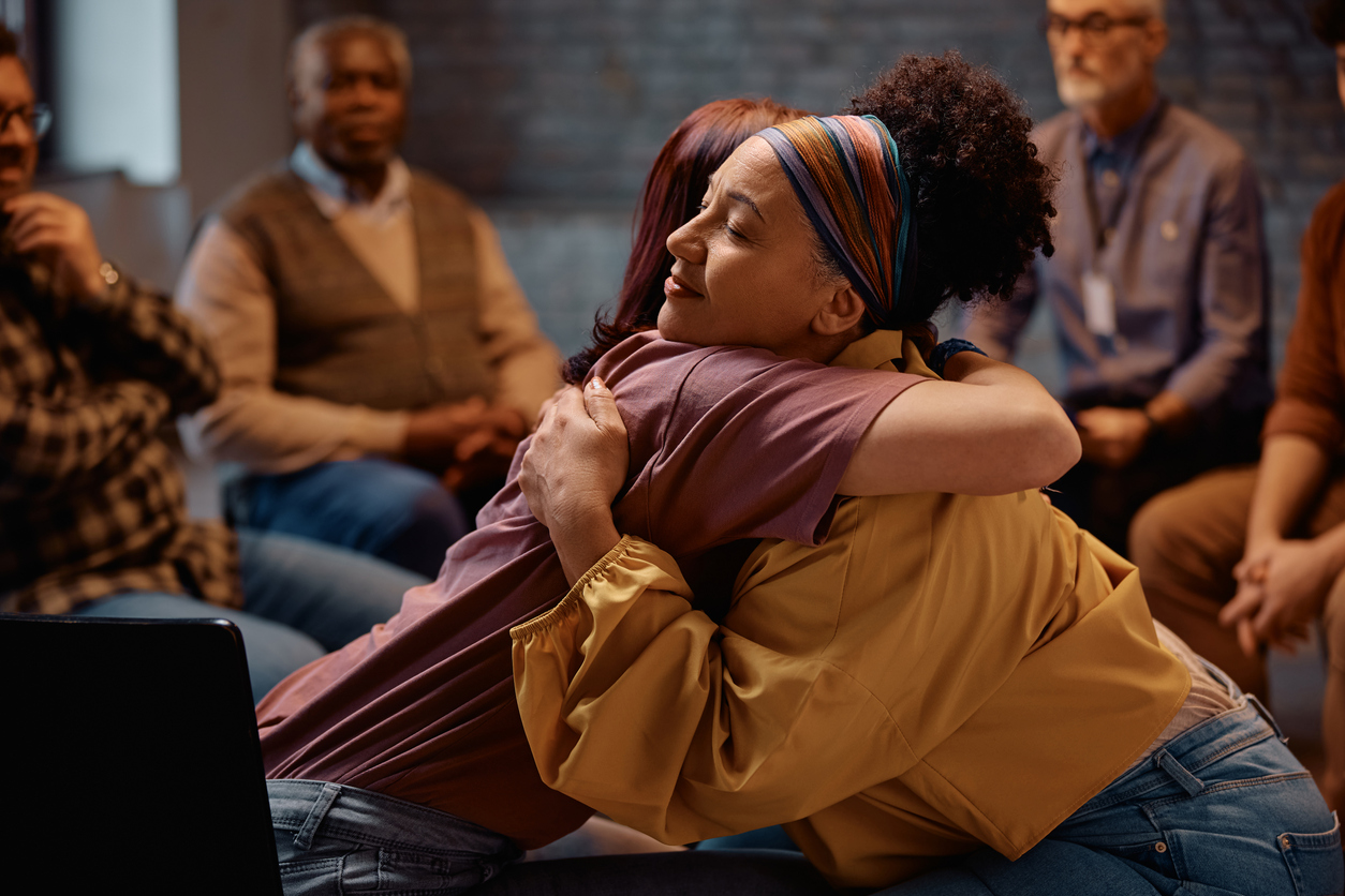 Supportive women embracing during group therapy at mental health center.
