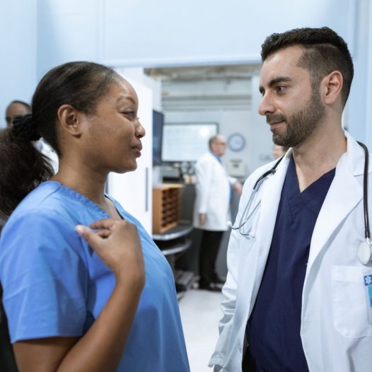 A Black registered nurse in blue scrubs talks with a clinician in a white coat in a busy hospital unit
