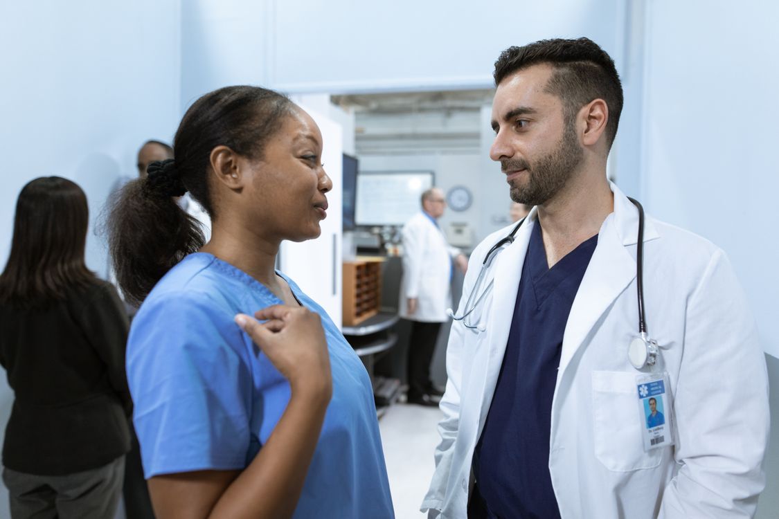A Black registered nurse in blue scrubs talks with a clinician in a white coat in a busy hospital unit